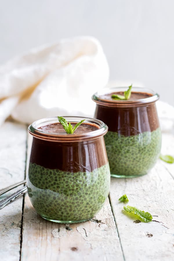 two jars of green chia pudding with chocolate smoothie on top on a white wooden table 