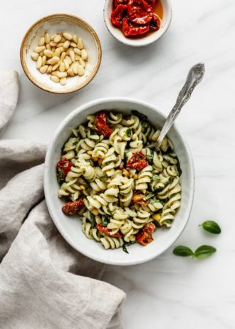 a bowl of pesto pasta salad with a napkin and some pine nuts on the side