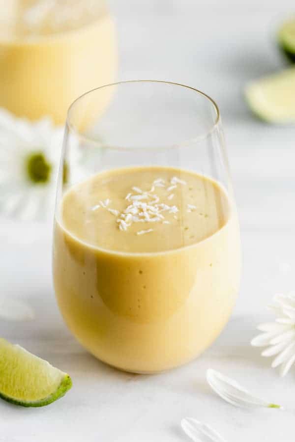 a glass of vegan mango lassi on a marble board with limes and flowers