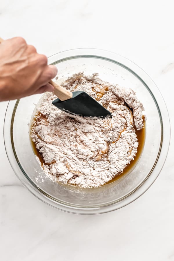 a mixing bowl with flour, eggs and sugar in it and a hand using a spatula to mix it together