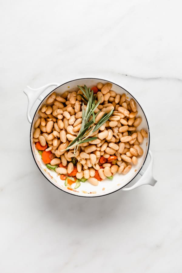 A white pot with white beans and a sprig of rosemary in it