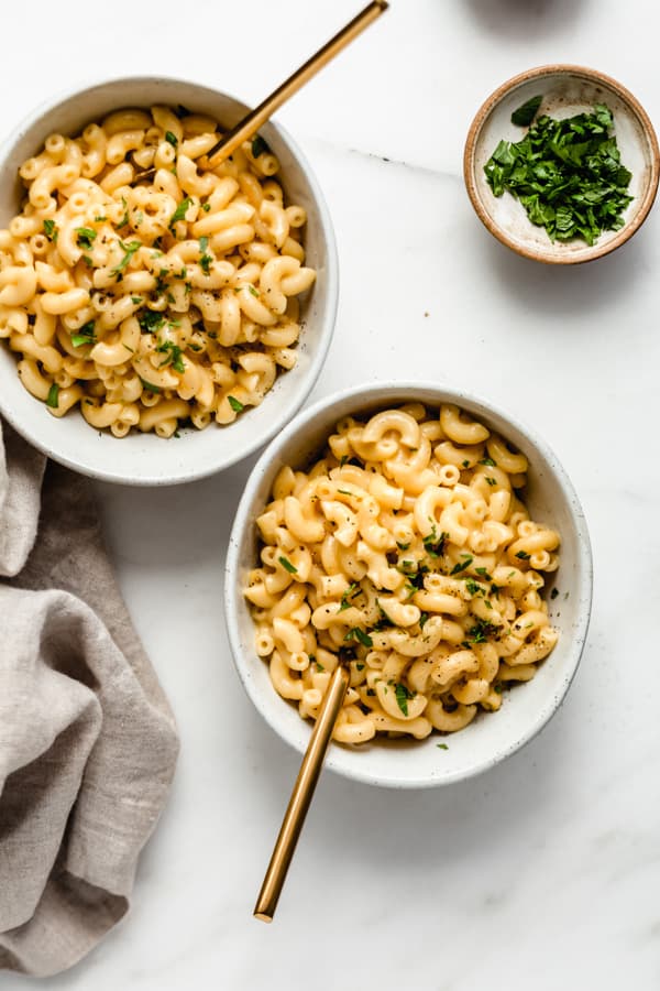 two bowls of mac and cheese on a marble board with a small bowl of parsley on the side