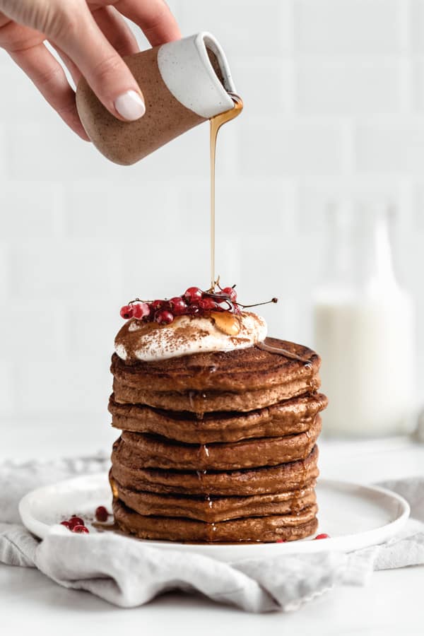a hand pouring maple syrup onto a stack of gingerbread pancakes