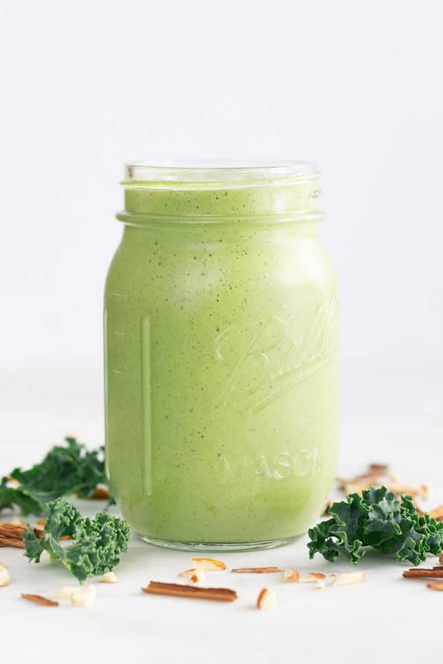 glass mason jar of healthy green breakfast smoothie surrounded by kale and almond slivers on a white background