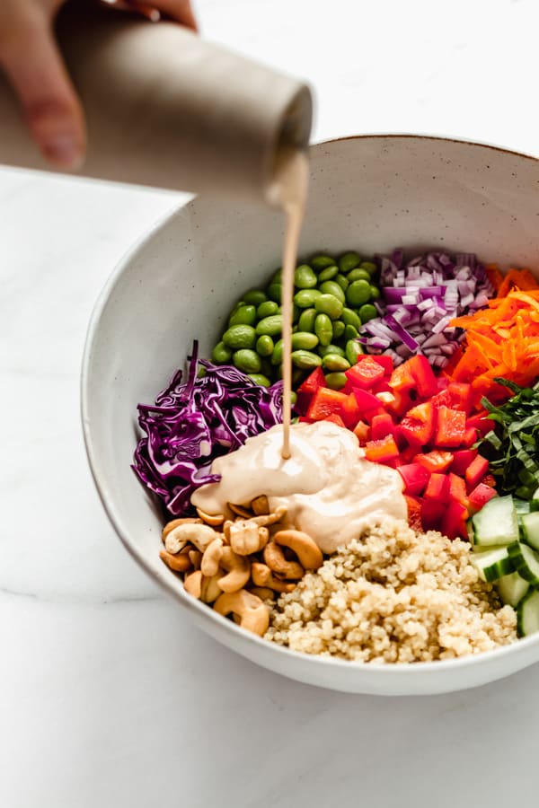 a hand pouring tahini salad dressing into a large mixing bowl filled with quinoa salad
