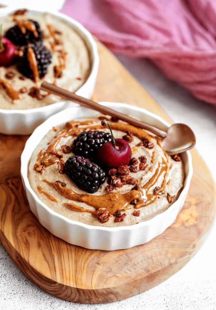 cherries and blackberries on top of a white ramekin filled with healthy breakfast pudding, drizzled with almond butter on top of a wooden board