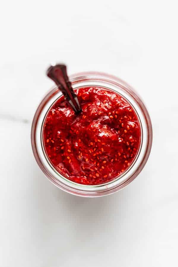 silver spoon sitting in a glass jar filled with strawberry chia jam on a marble counter