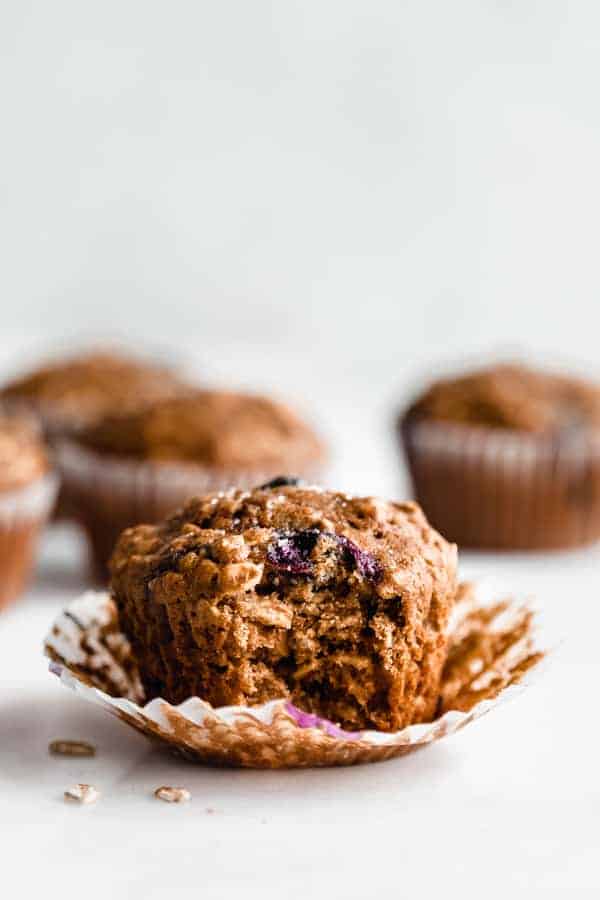 a healthy breakfast muffin with a bite taken out of it, on the muffin paper with blueberry muffins in the background