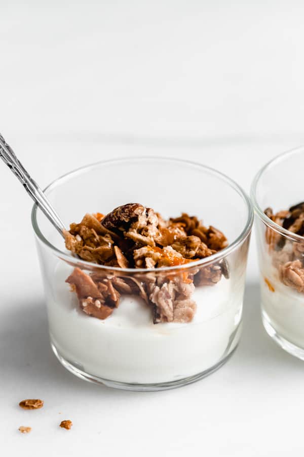a silver spoon in a glass jar of yogurt topped with orange granola against a white background