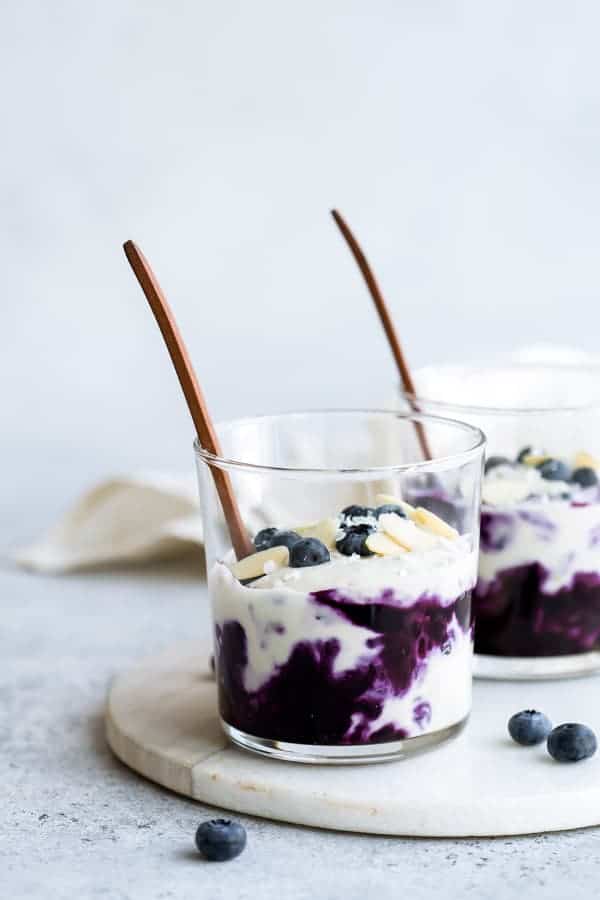 jars of blueberry cheesecake with wooden spoons on top, on top of a wooden board with blueberries and a white napkin