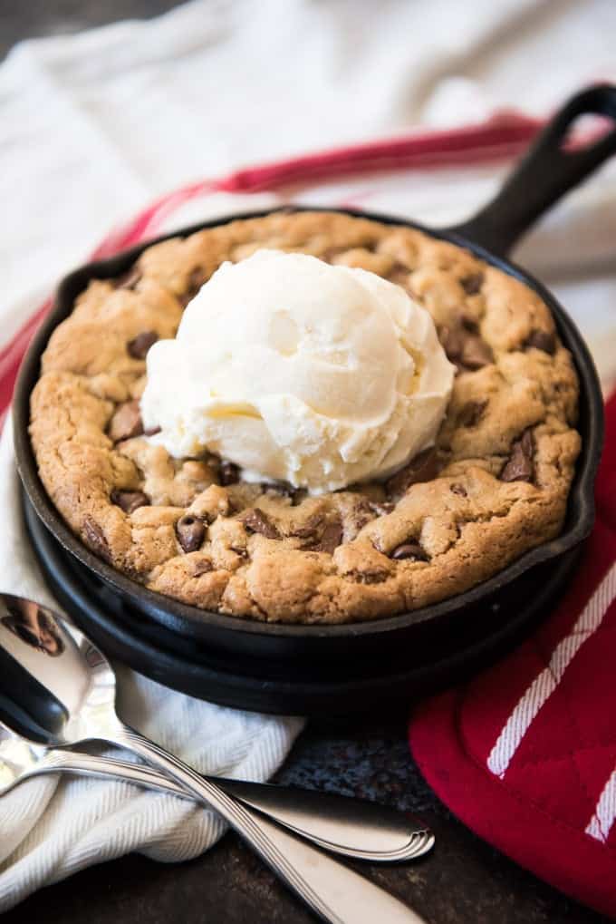 one cast iron chocolate chip cookie skillet with a scoop of vanilla ice cream and two silver spoons next to the skillet, on top of a red oven mitt and white linen on a black countertop