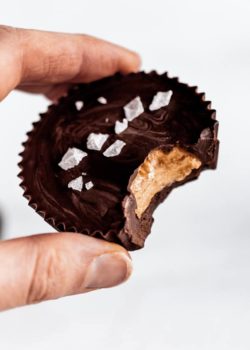 A hand holding a dark chocolate almond butter cup with a bite taken out of it