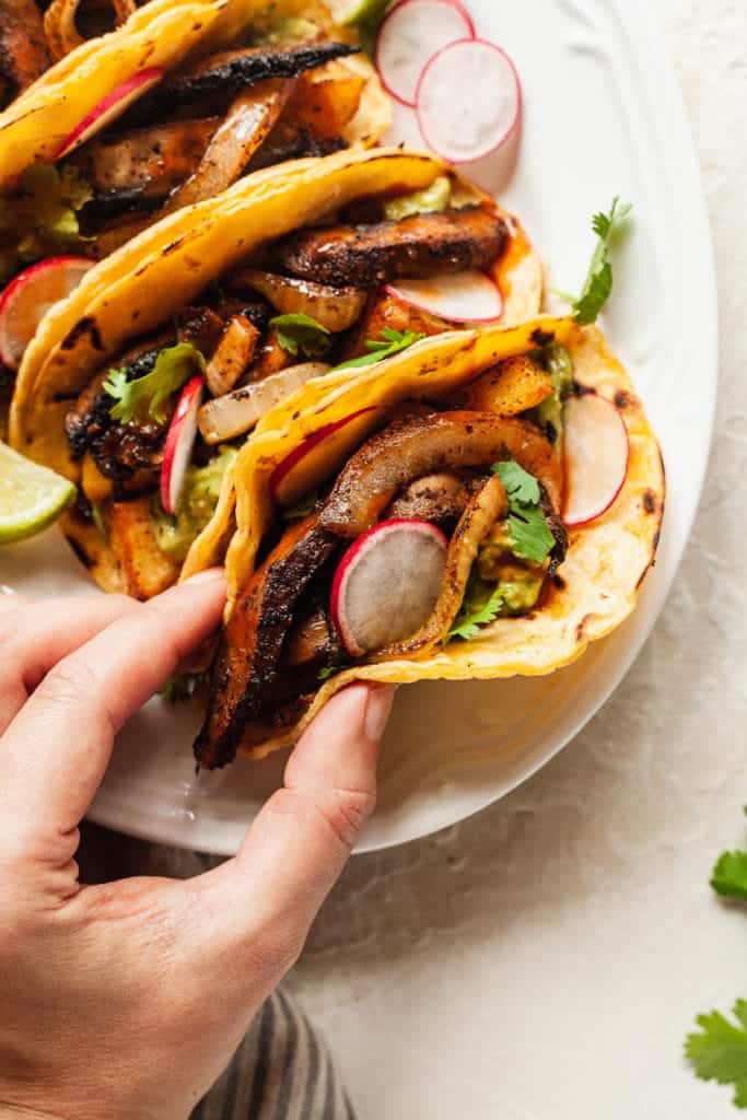 hand reaching for a taco filled with vegetables, cilantro, and sliced radishes lined up on a white serving platter with lime wedges for date night