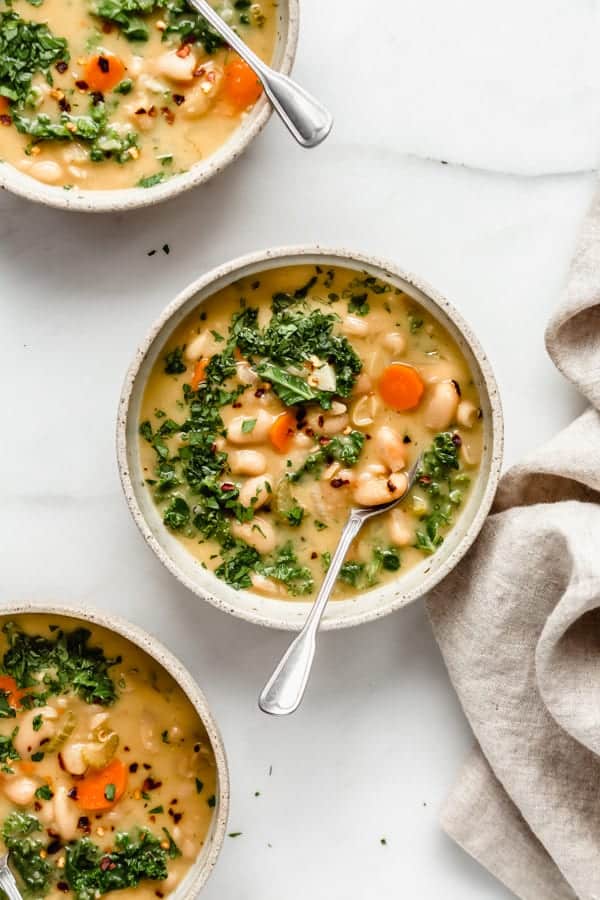 white bean soup with kale and carrots and red chili pepper flakes in tan bowls with a tan linen on a marble countertop