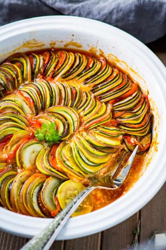 sliced vegetables arranged in concentric circles and covered in tomato sauce to make ratatouille, with a silver serving fork in the middle of the white dish 