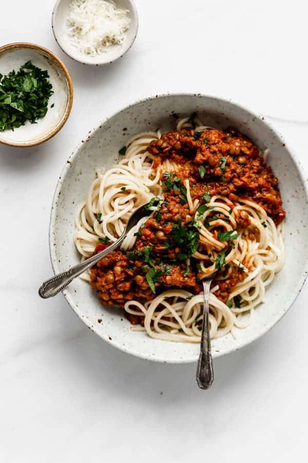 two silver spoons in a bowl of spaghetti pasta topped with bolognese and herbs in a ceramic bowl, with two pinch bowls filled with herbs and cheese next to the bowl on a white table
