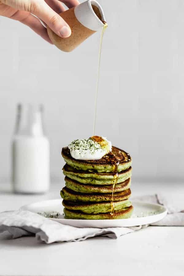 a hand pouring syrup onto a stack of matcha pancakes