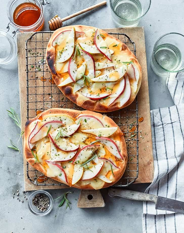 pear, brie, and rosemary flatbreads for date night on a wooden platter surrounded by fresh herbs on a blue backdrop with a silver knife