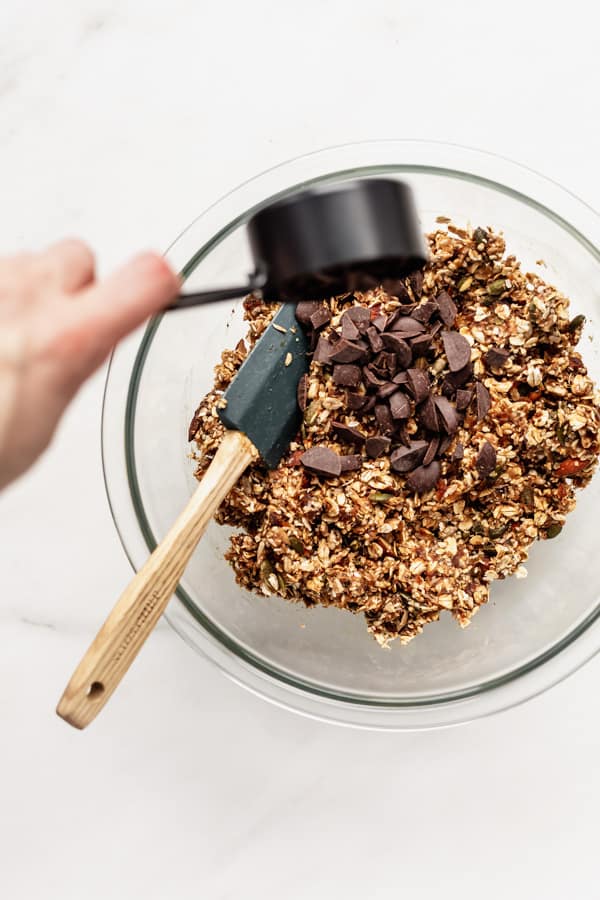 a hand pouring chocolate chunks into a bowl of superfood granola bars ingredients