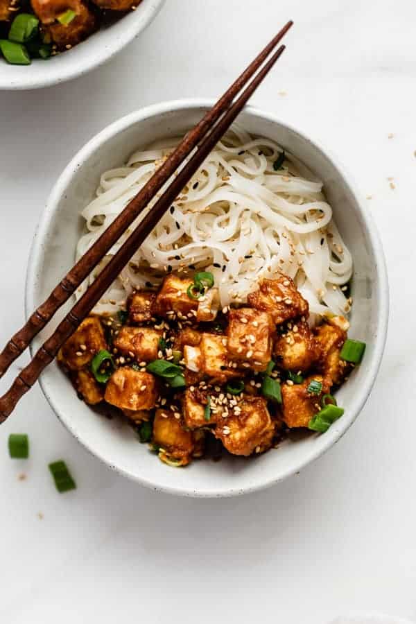 a bowl of noodles topped with peanut butte tofu, sesame seeds and green onions