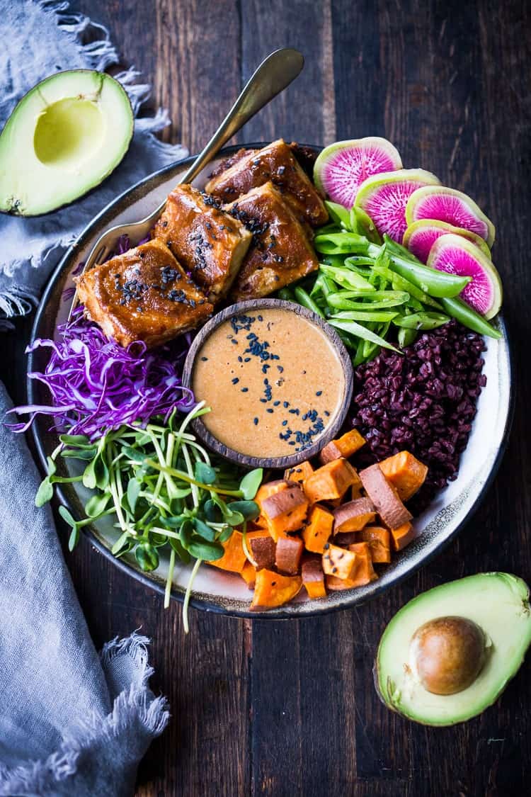 peanut sauce in a wooden bowl in the middle of a ceramic bowl filled with peanut tofu, cabbage and microgreens and sweet potatoes