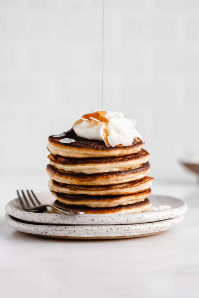 a stack of coconut flour pancakes topped with yogurt and coconut flakes with maple syrup being drizzled on top