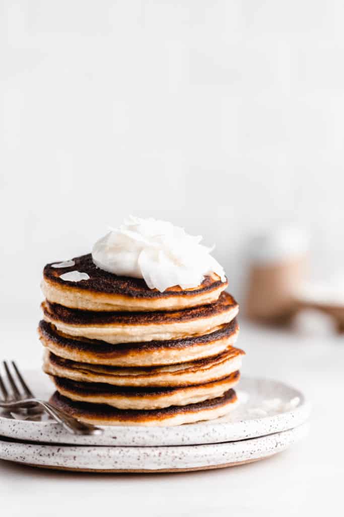 a stack of coconut flour pancakes topped with yogurt and coconut flakes with maple syrup being drizzled on top