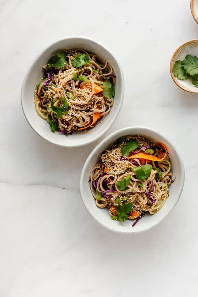 two bowls of soba noodle salad topped with sesame seeds, green onions and cilantro