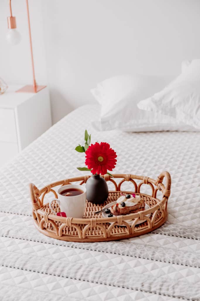 A rattan trey with a flower, tea and french toast on it on a bed 