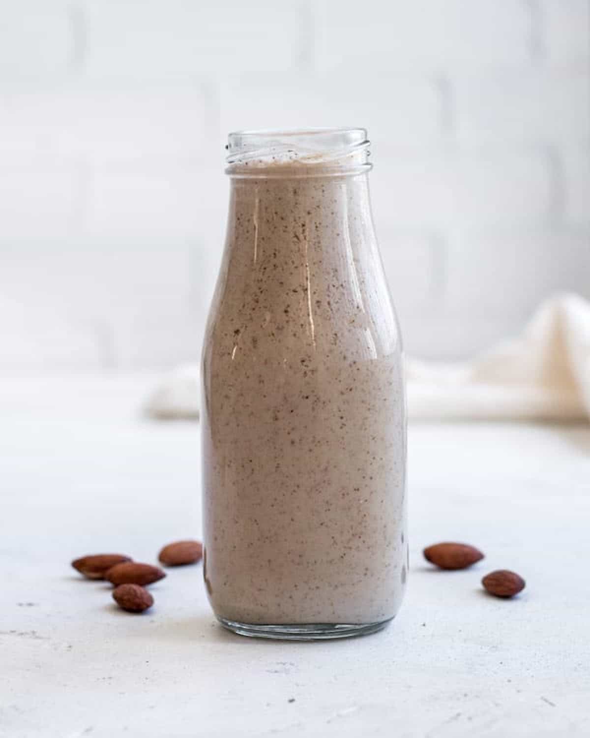 one brown speckled smoothie in a milk jar surrounded by whole almonds on a white background