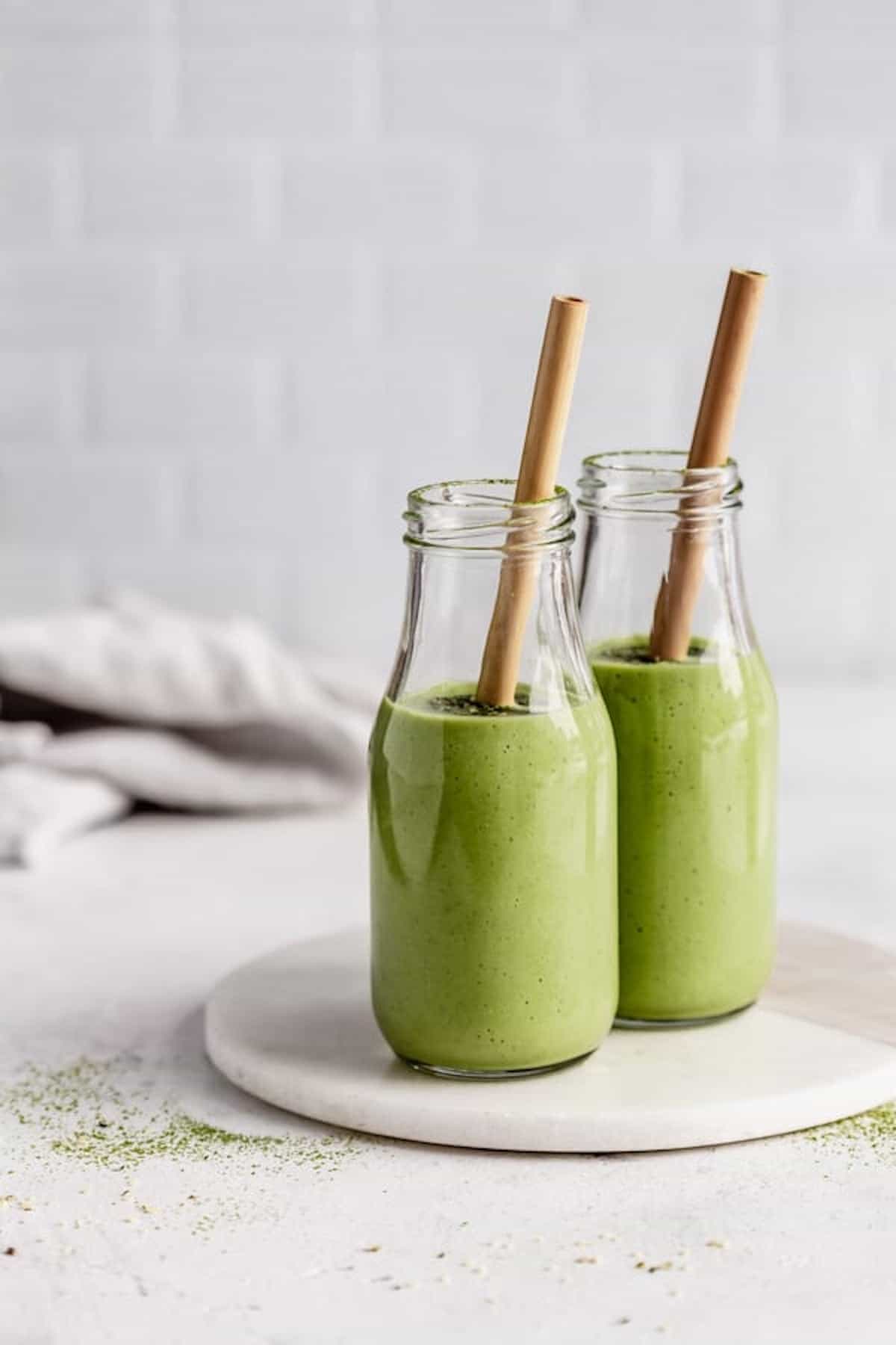 two brown paper straws sticking out of two tall glasses of matcha green tea smoothie on top of a marble serving platter against a white backdrop, healthy smoothie