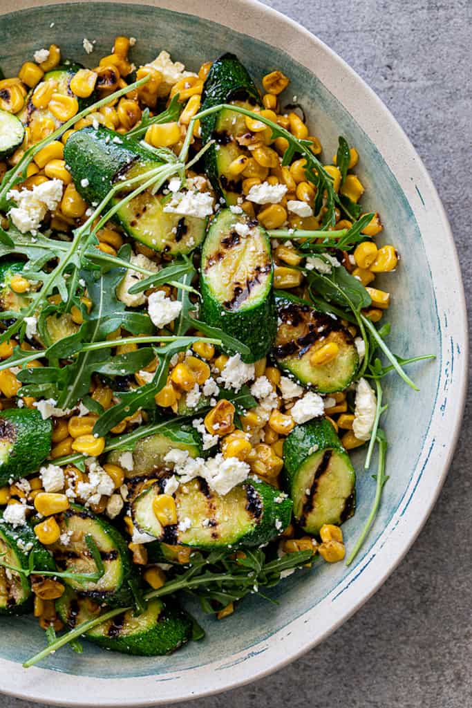 grilled summer salads made of grilled zucchini and corn niblets mixed with crumbled feta cheese and fresh green arugula, in a white and blue ceramic bowl, against a white background