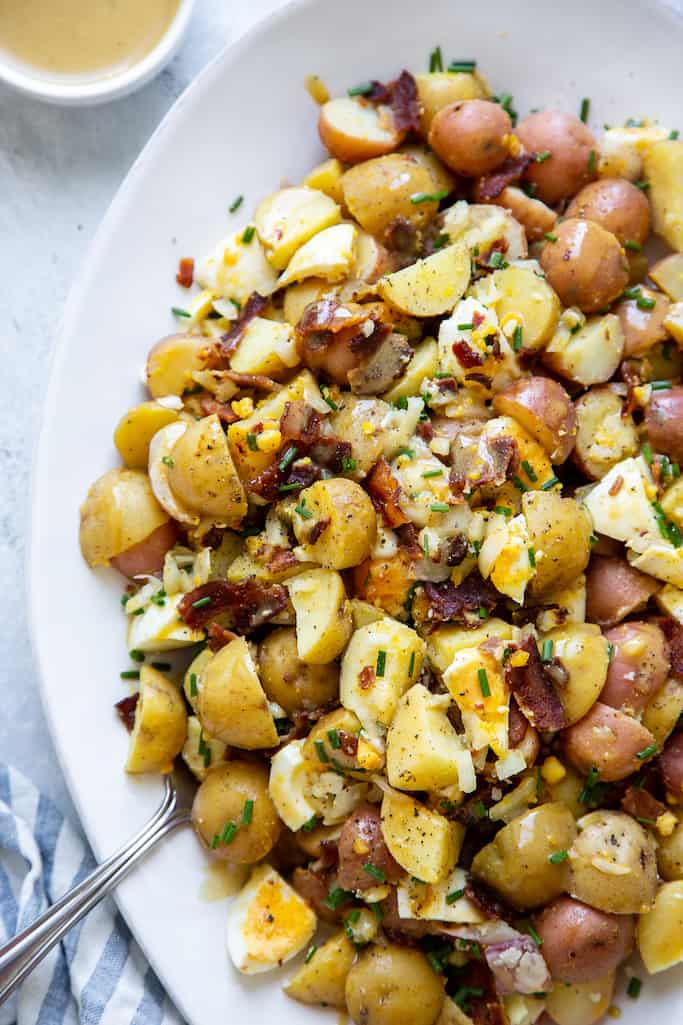 honey dijon potato salad mixed with boiled eggs, bacon, chives, and a honey mustard dressing on a white serving platter