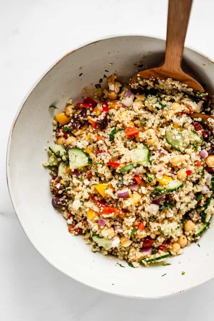 white ceramic bowl filled with summer salads made of quinoa, cucumbers, red and yellow peppers, with a large wooden spoon