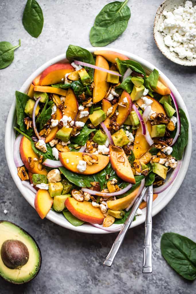 summer peach spinach salad with sliced peaches, red onions and toasted almonds in a large white bowl with chopped avocado and silver utensils, against a grey background