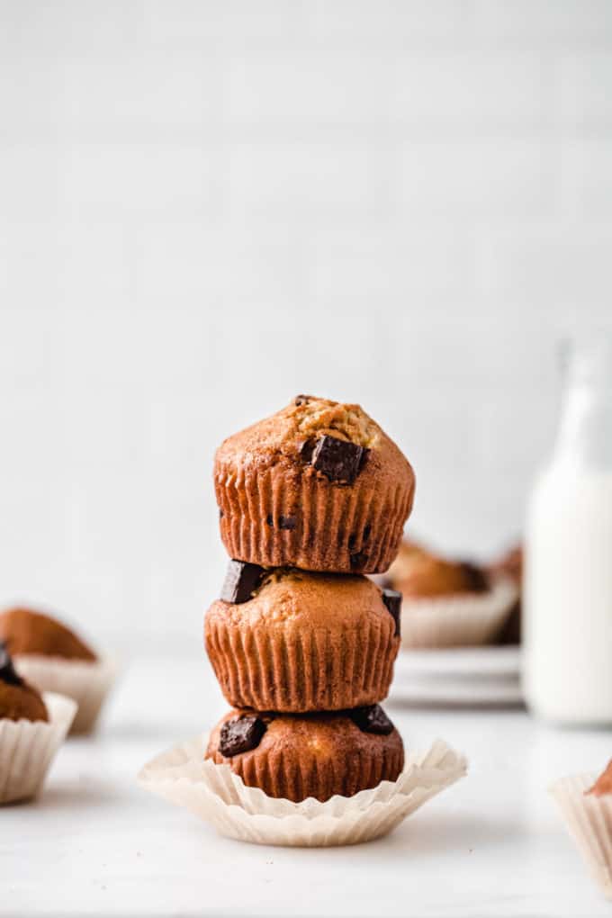 three chocolate chip banana muffins stacked on top of each other with a jar of milk in the background