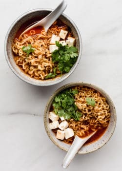 two white speckeld ceramic bowls of kimchi ramen with spoons in them