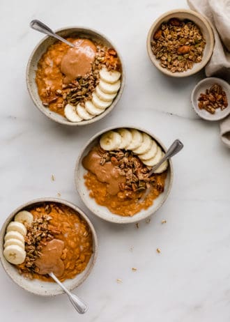 three bowls of pumpkin pie oatmeal with small bowls of granola and pecans on the side