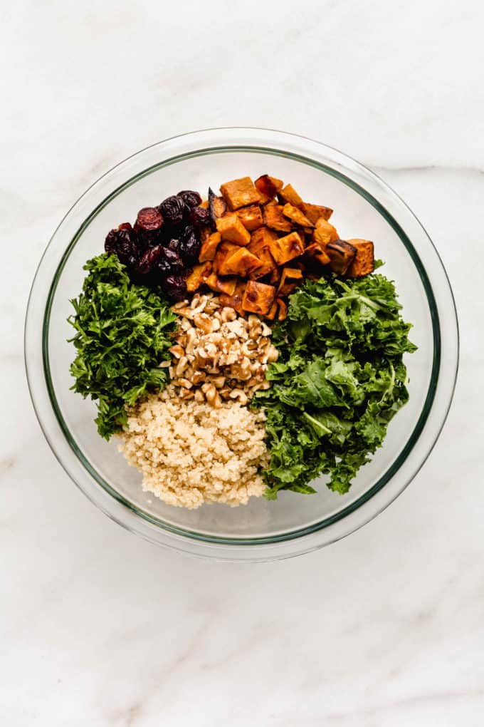A clear mixing bowl with kale, quinoa, parsley, sweet potatoes, cranberries and walnuts in it