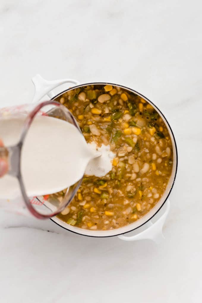 A measuring cup pouring cashew cream into a pot of white chili