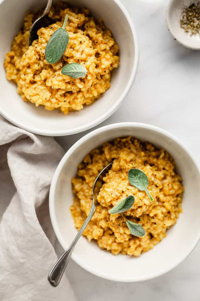 Two bowls of butternut squash risotto topped with sage with a pinch bowl of pepper on the side