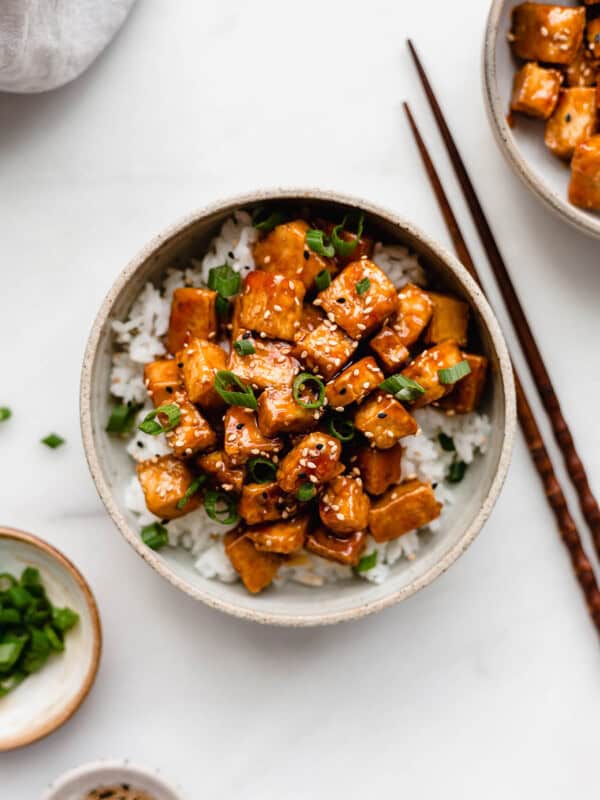 Sweet and sour tofu in a white ceramic bowl with two small bowls on the side filled with green onions and sesame seeds