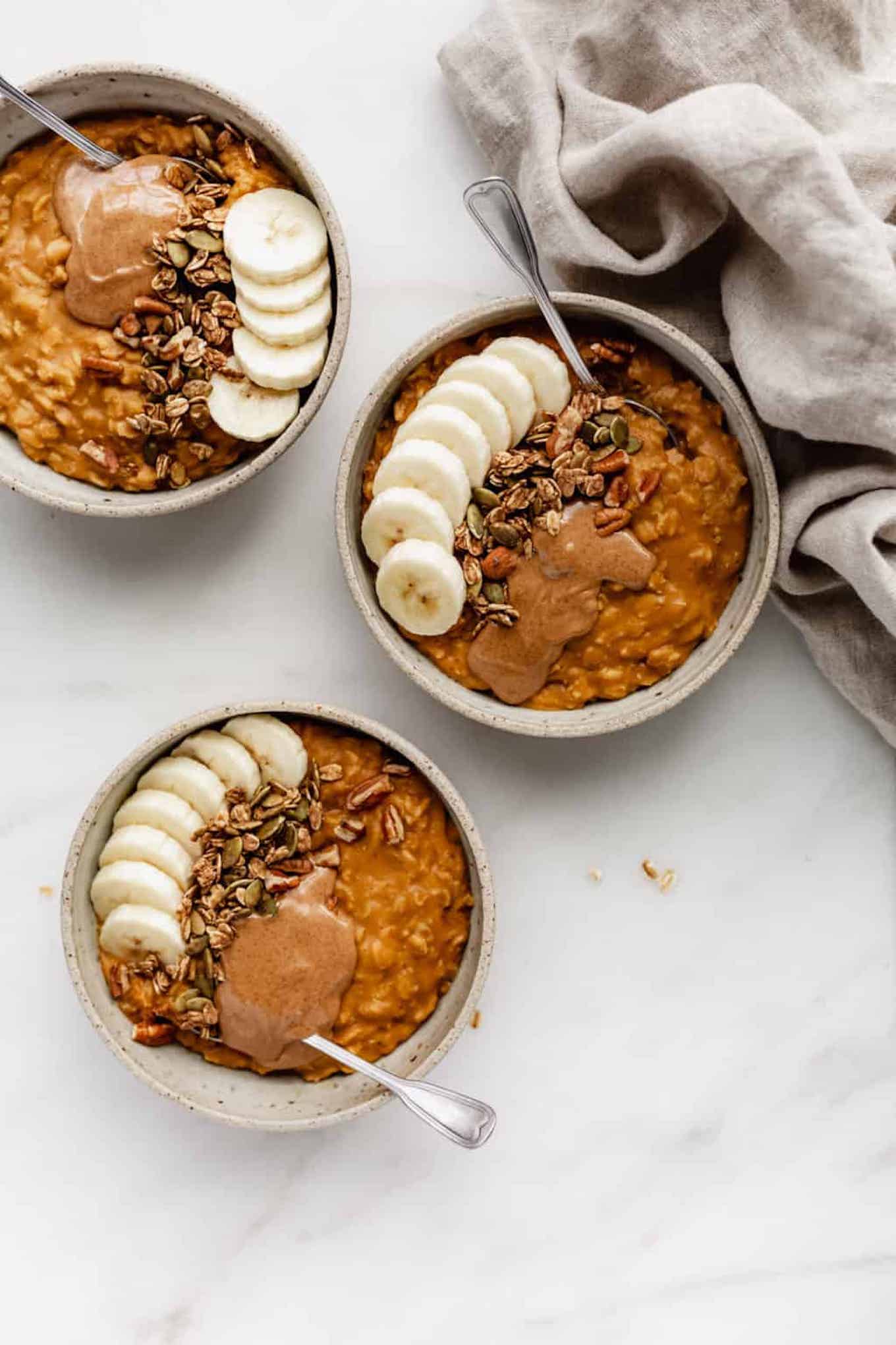 three bowls of pumpkin pie oatmeal topped with sliced bananas, granola, and pecans