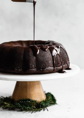 A gingerbread bundt cake on a marble cake stand with chocolate glaze being poured onto it