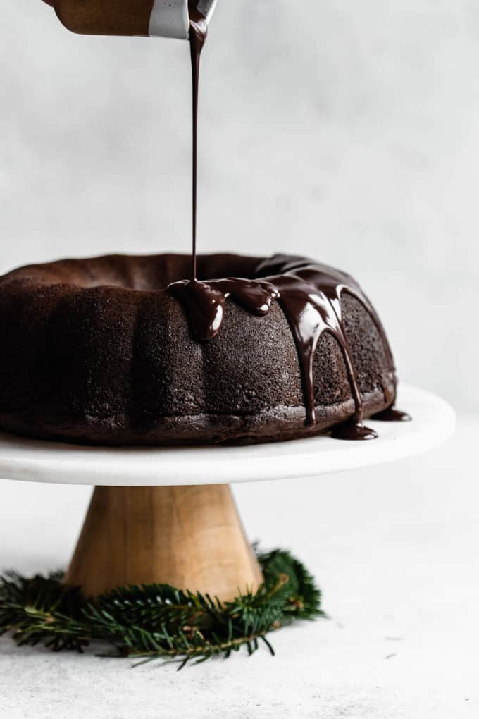 A gingerbread bundt cake on a marble cake stand with chocolate glaze being poured onto it