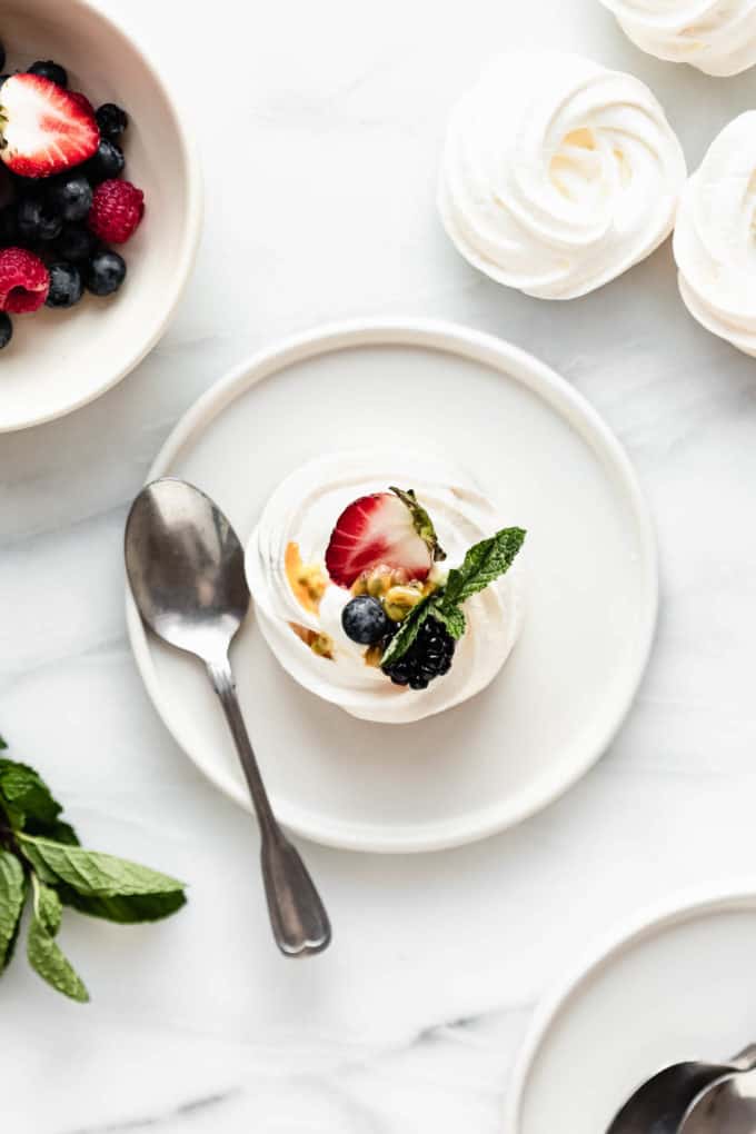 A plate with a spoon and a mini pavlova on it with meringues and berries on the side