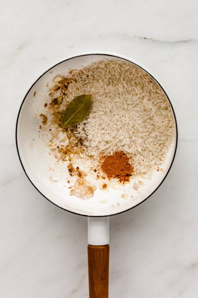 A white pot with shallots, turmeric, rice and a bay leaf in it