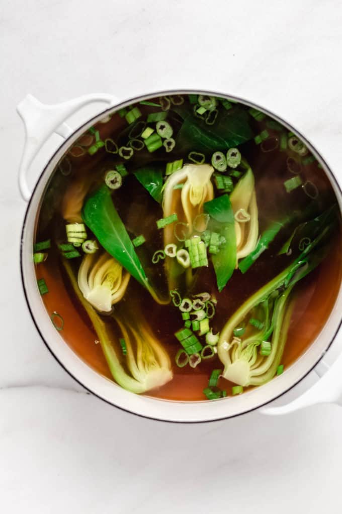 Vegan pho broth in a white pot with green onions and bok choy
