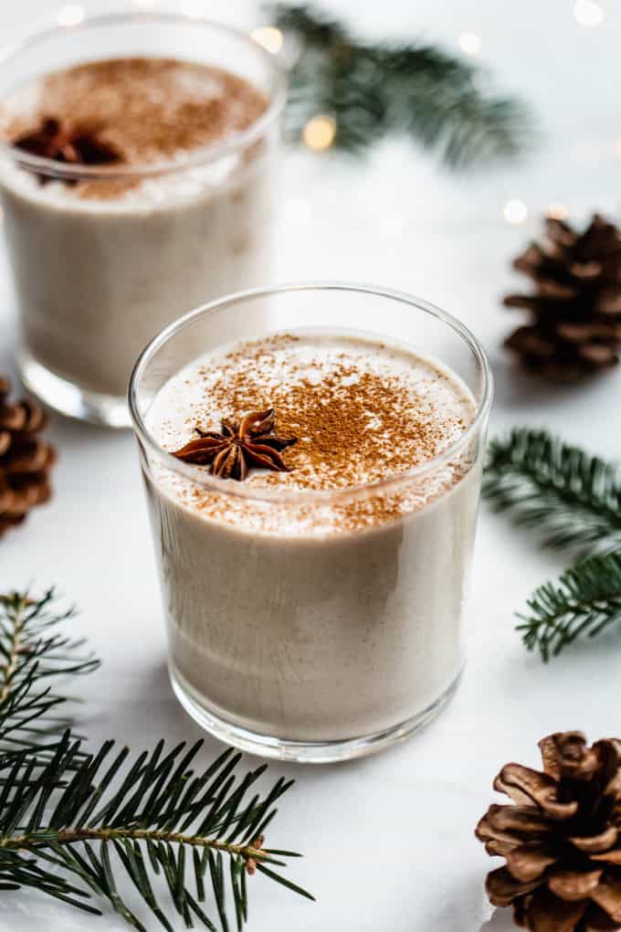 A glass of vegan eggnog on a white counter surrounded by pinecones and pine leaves