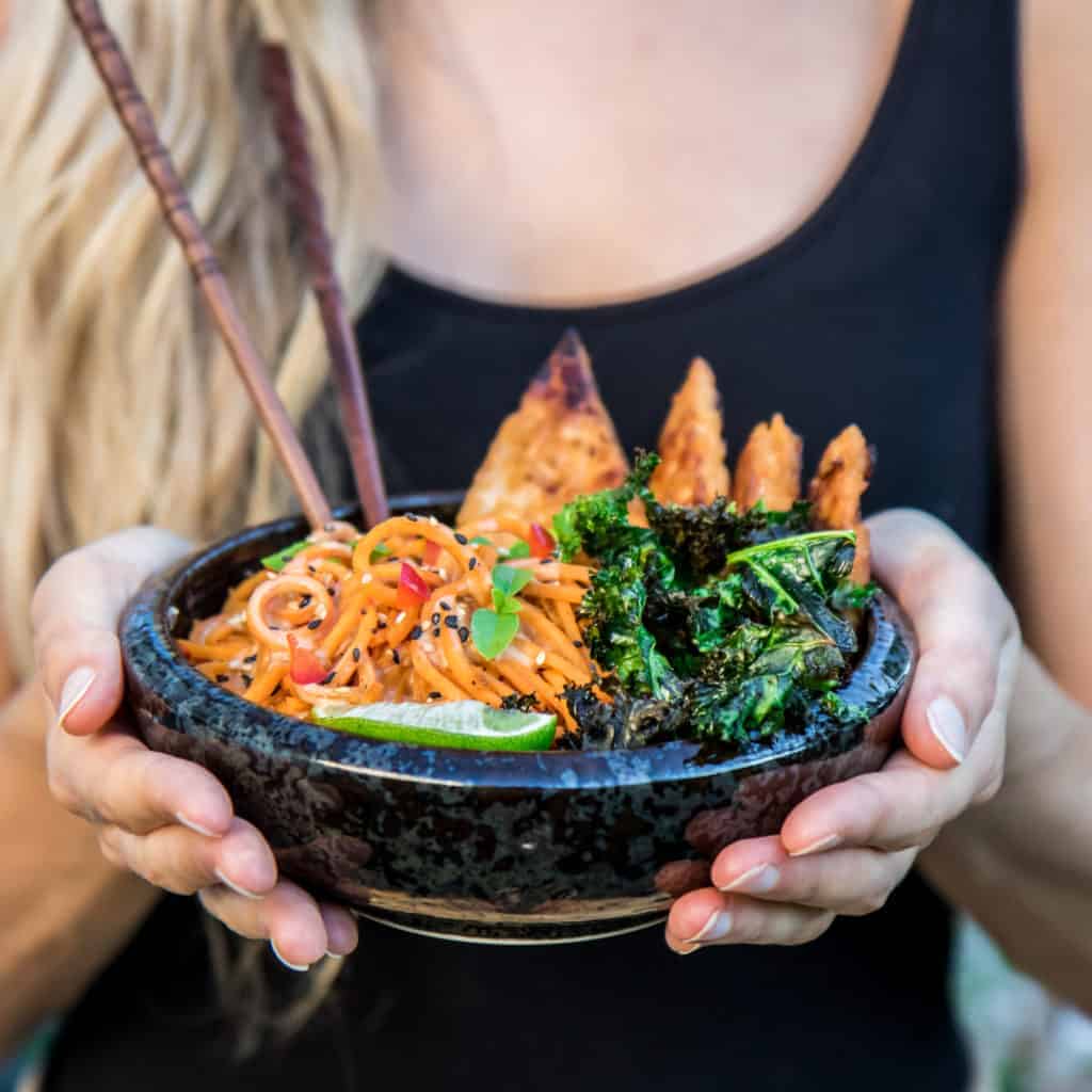 A person holding a black bowl with sweet potato noodles and tempeh in it
